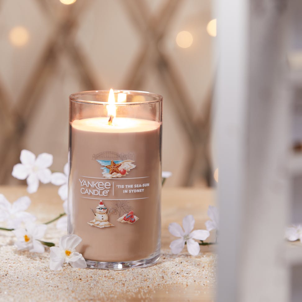 Passport to the Holidays Collection Tis the Sea-Sun in Sydney Signature Medium Pillar Candle on sand surrounded by white flowers