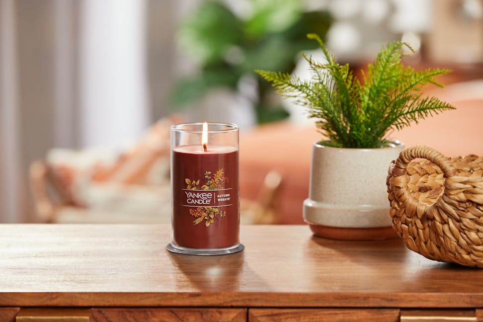 lit autumn wreath signature medium pillar candle on wooden table next to a potted plant and basket