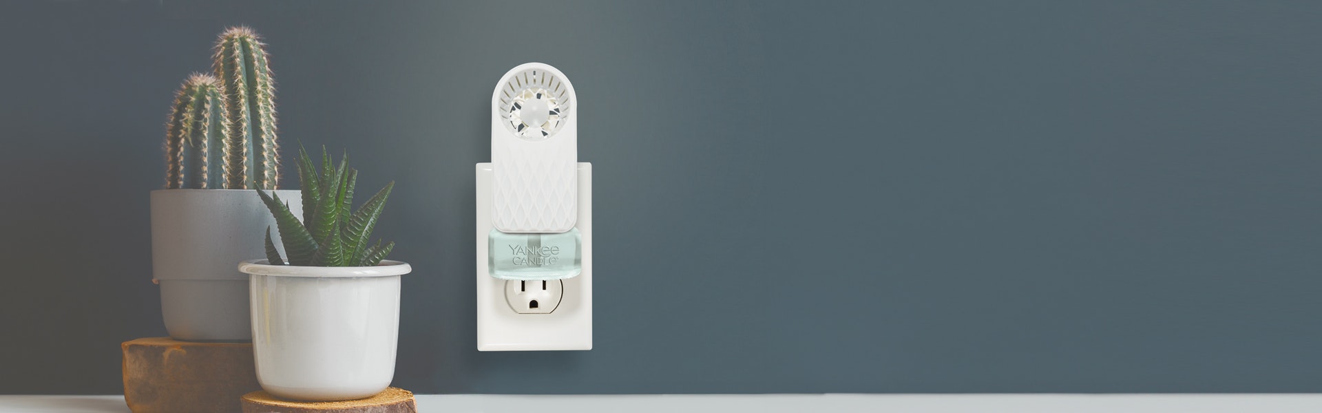 Fade ScentPlug Fan with a light blue-colored ScentPlug Refill plugged into a socket next to two plants