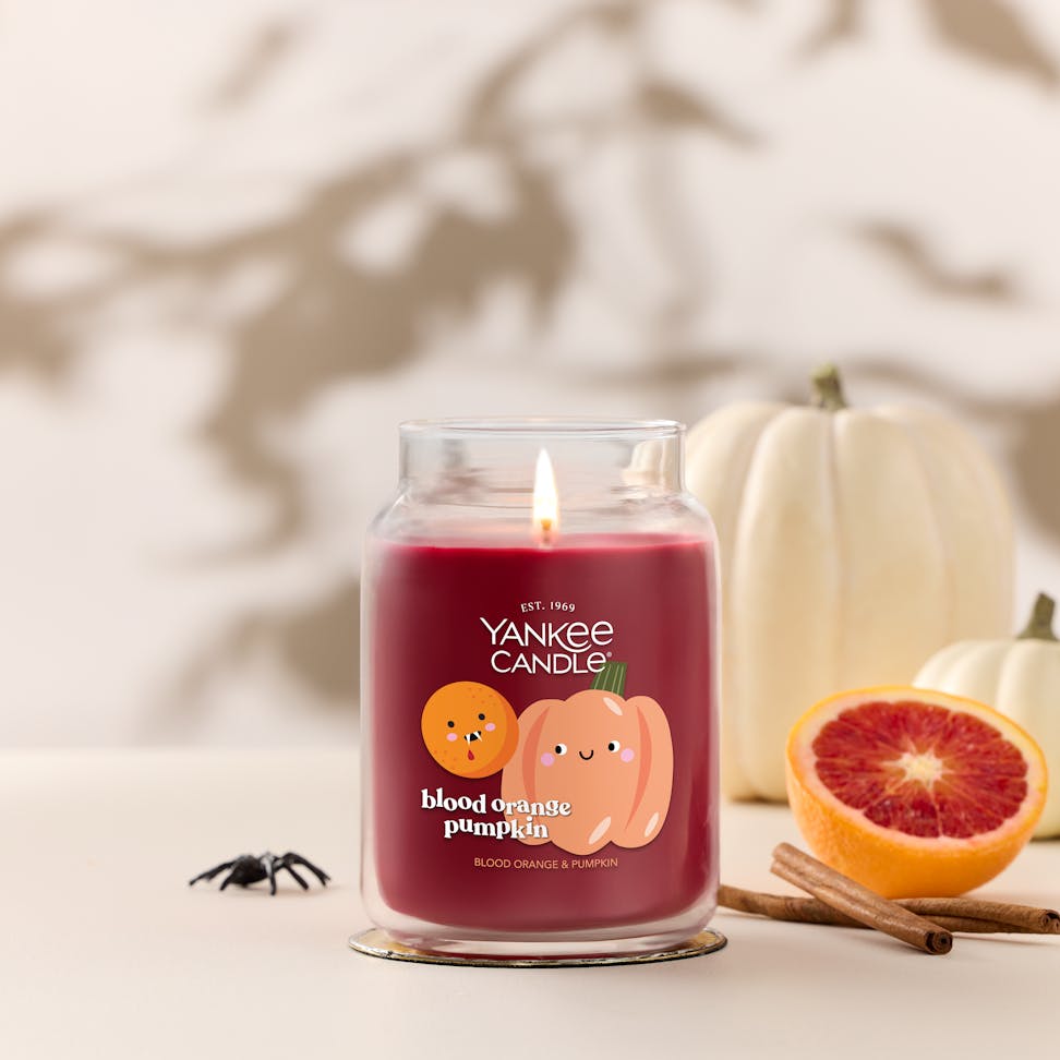 Large red pumpkin candle with cute fruit graphics, lit beside pumpkin, orange, cinnamon and a small black spider.