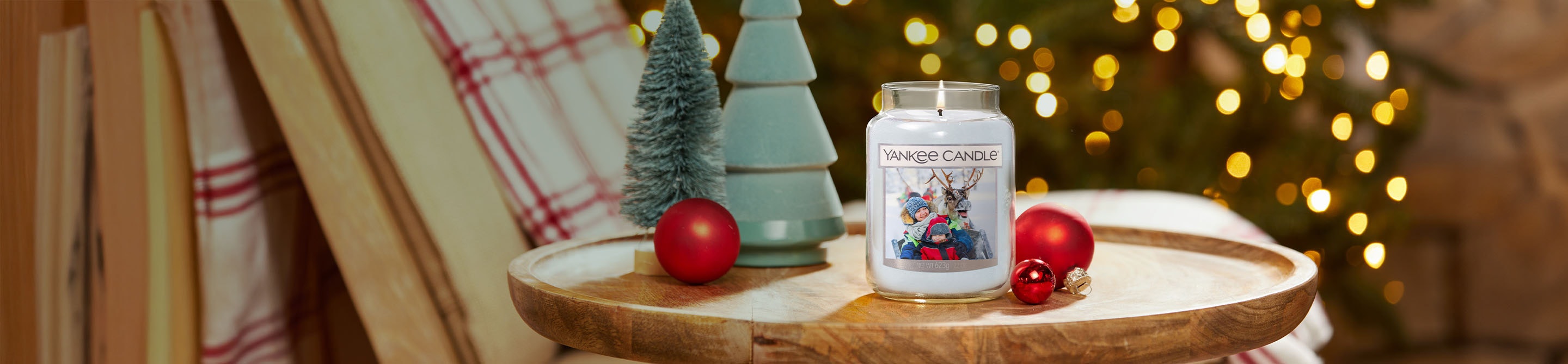 Large holiday candle jar with festive label on wooden tray, surrounded by Christmas decor and ornaments