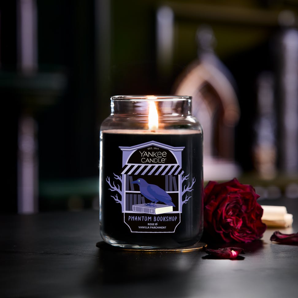 Large black scented candle in glass jar with a raven label, burning beside a red rose and stacked books