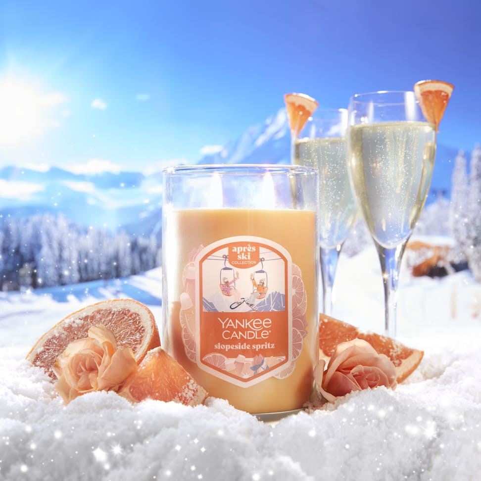 large tumbler candle in slopeside spritz in snowy mountain landscape with sliced grapefruit and roses and glasses of sparkling wine