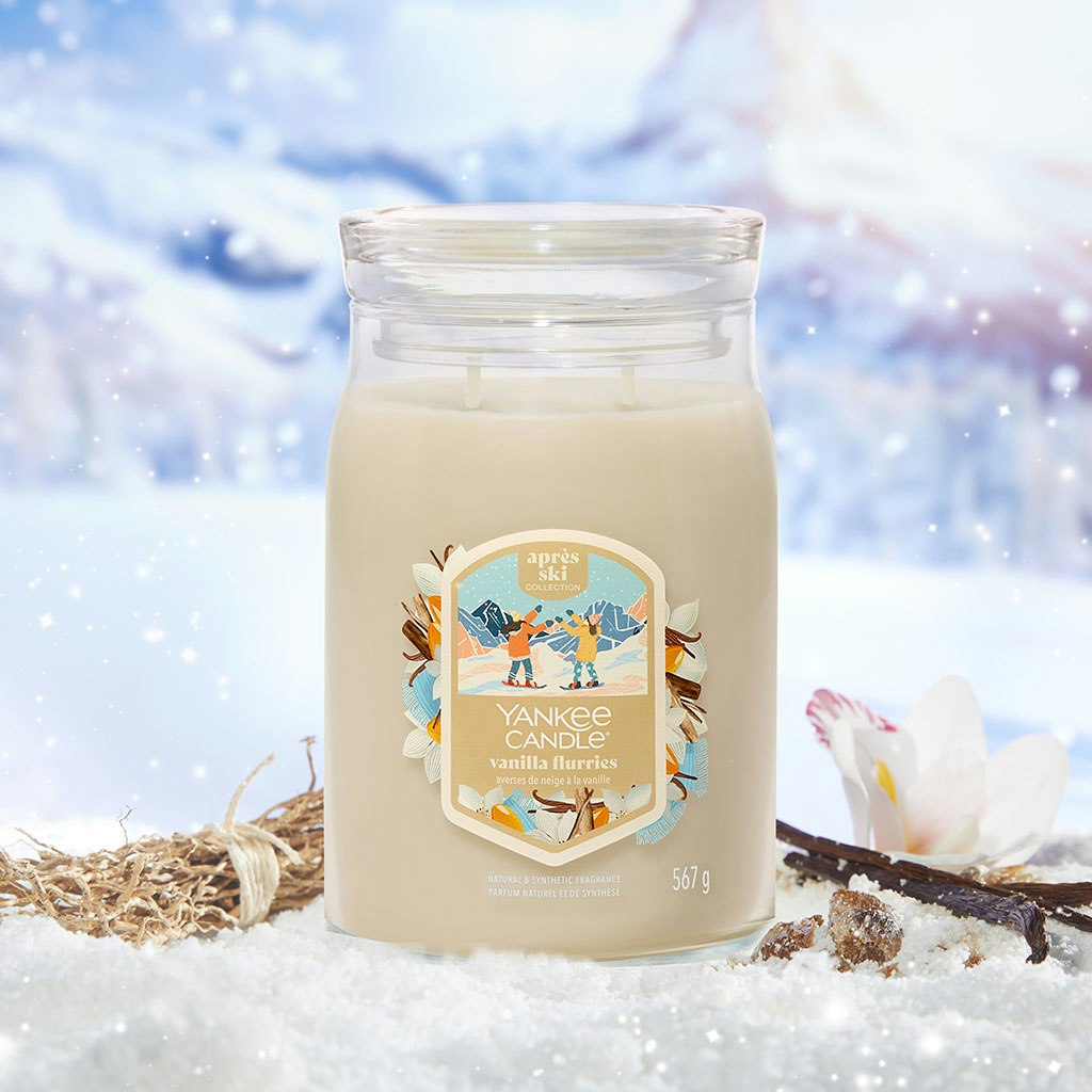 Large cream-colored scented candle jar with winter-themed label, surrounded by snow, flower, and vanilla pods.