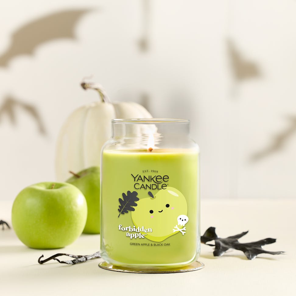 Large green apple scented candle in glass jar with autumn leaves, pumpkin, apple, and Halloween decor in background