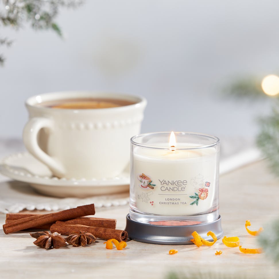 Passport to the Holidays Collection London Christmas Tea Signature Small Tumbler Candle on a table next to a white teacup and plate set and cinnamon sticks