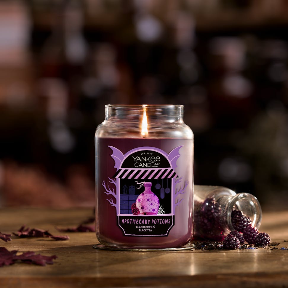 Large purple jar candle with apothecary label lit on rustic table beside spilled decorative blackberries