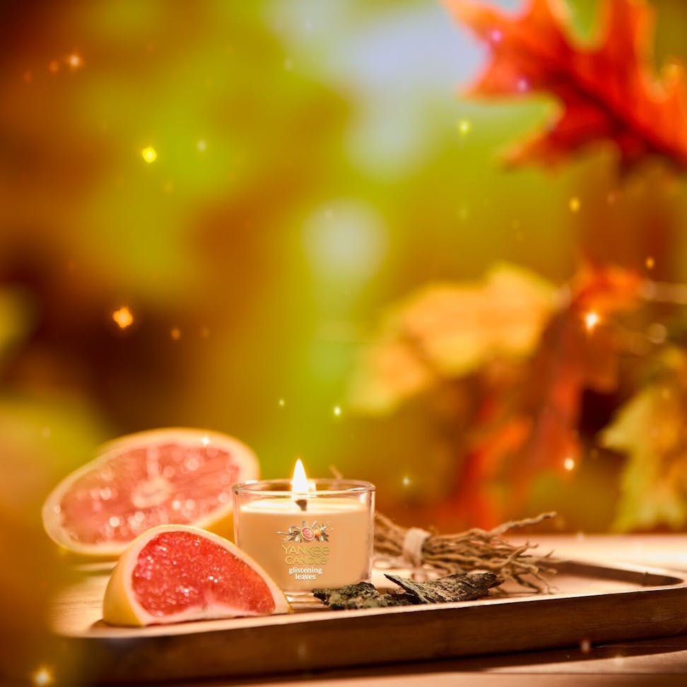 Lit glass candle with grapefruit slices and sage on a tray, autumn leaves and glowing lights in the background