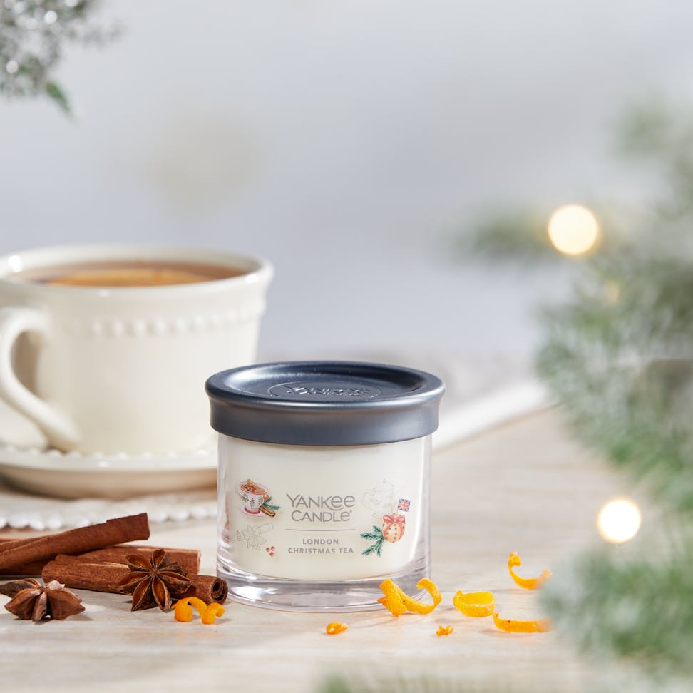 Passport to the Holidays Collection London Christmas Tea Signature Small Tumbler Candle on a table next to a white teacup and plate set and cinnamon sticks