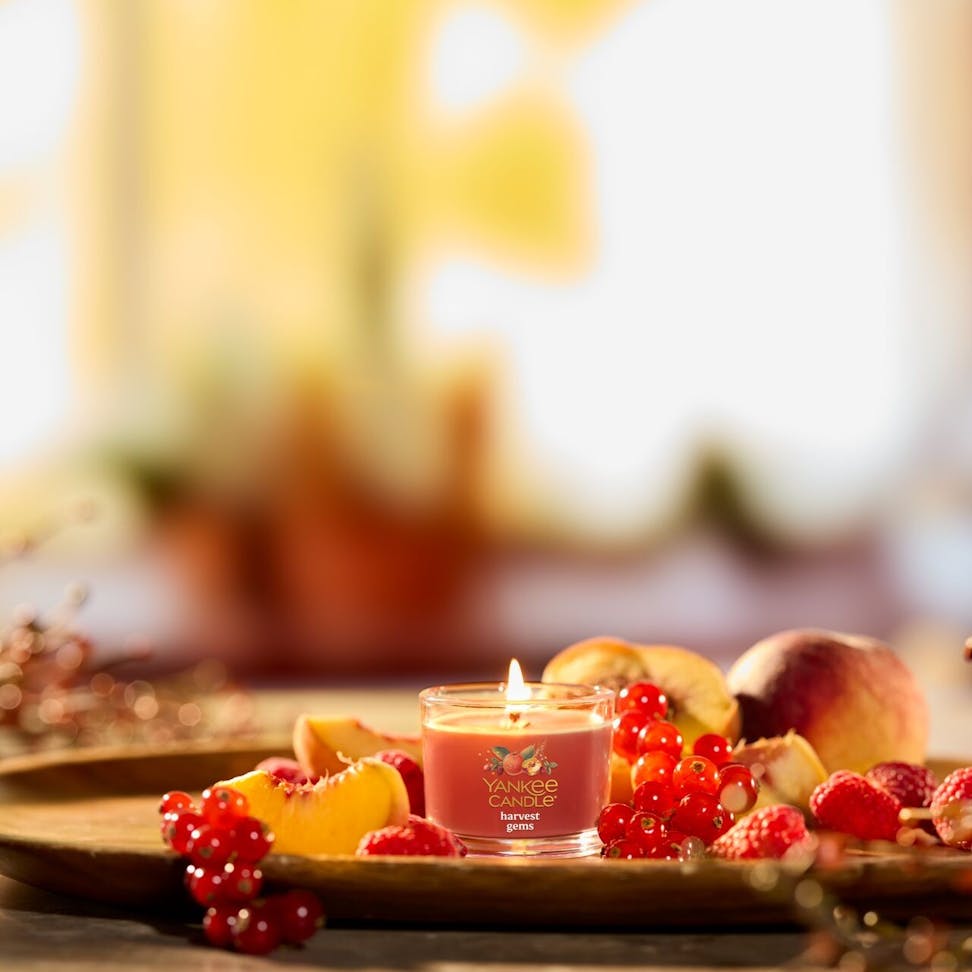 Scented candle in glass jar surrounded by fresh berries and peaches on a wooden tray for home decor.