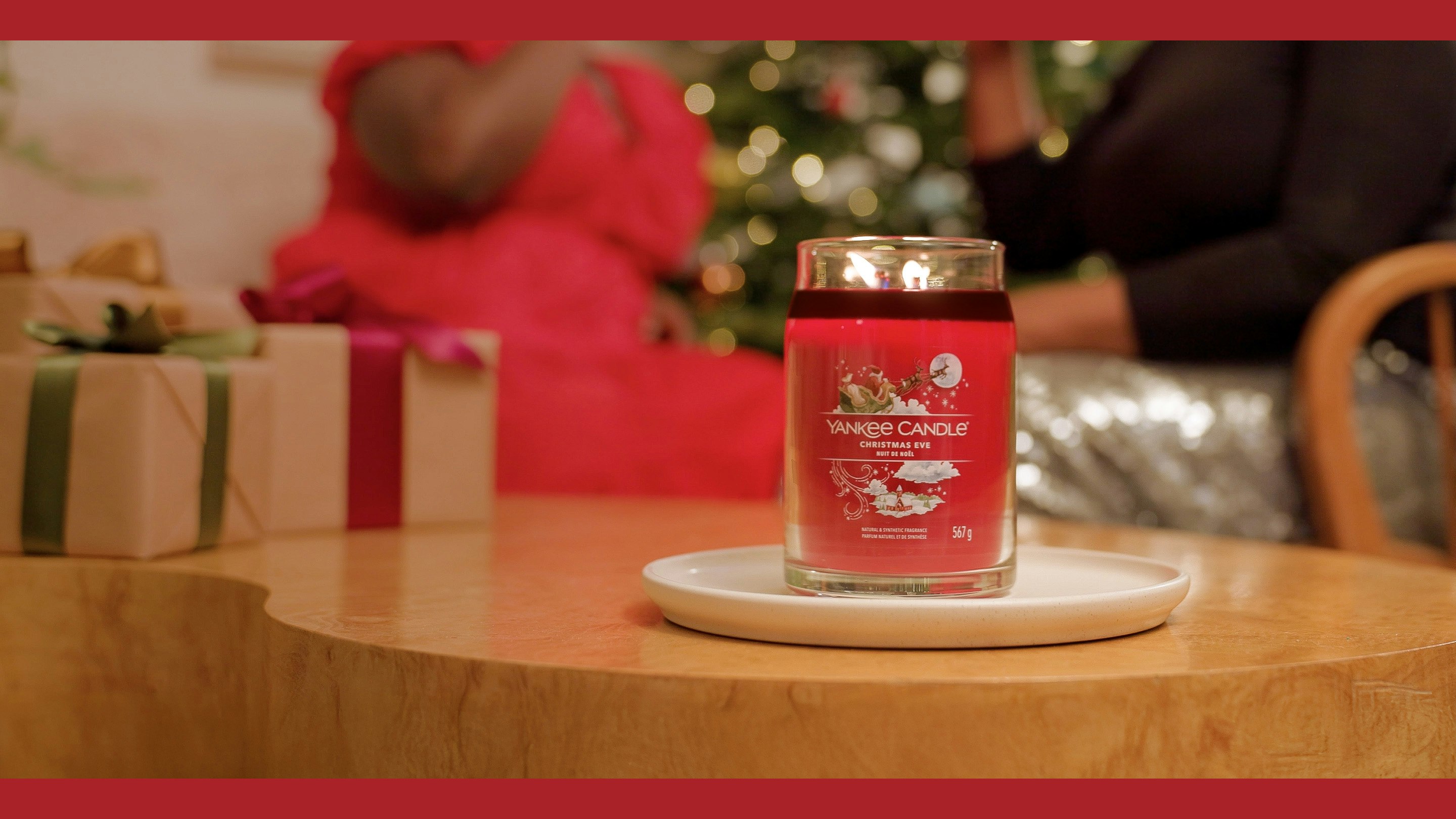 Large holiday candle jar with festive label on a table, Christmas tree and gift box in cozy living room setting