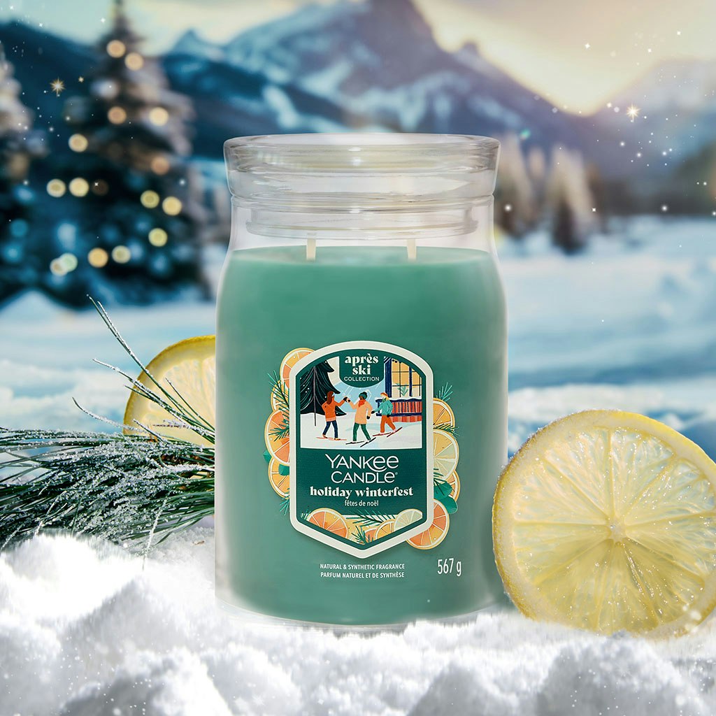 Large green holiday candle with winter scene label, surrounded by pine and lemon, snowy landscape in background