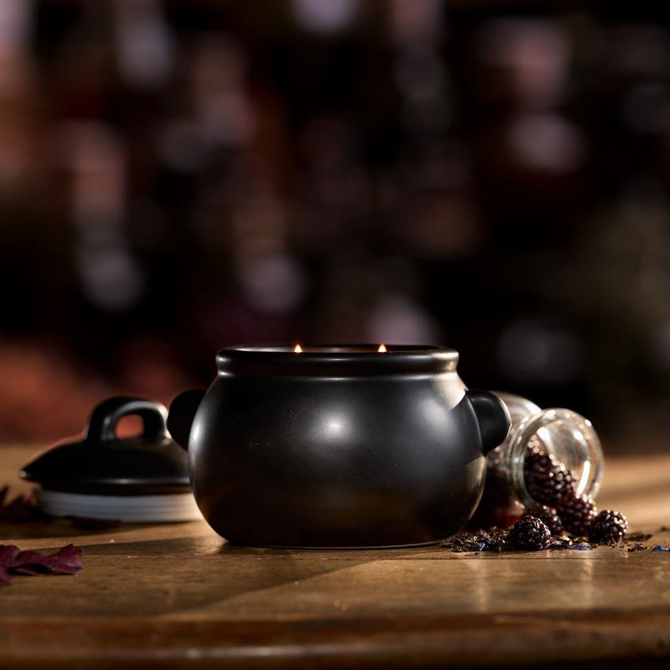 Black ceramic cauldron candle with two wicks on wooden table, dried berries and petals scattered nearby.
