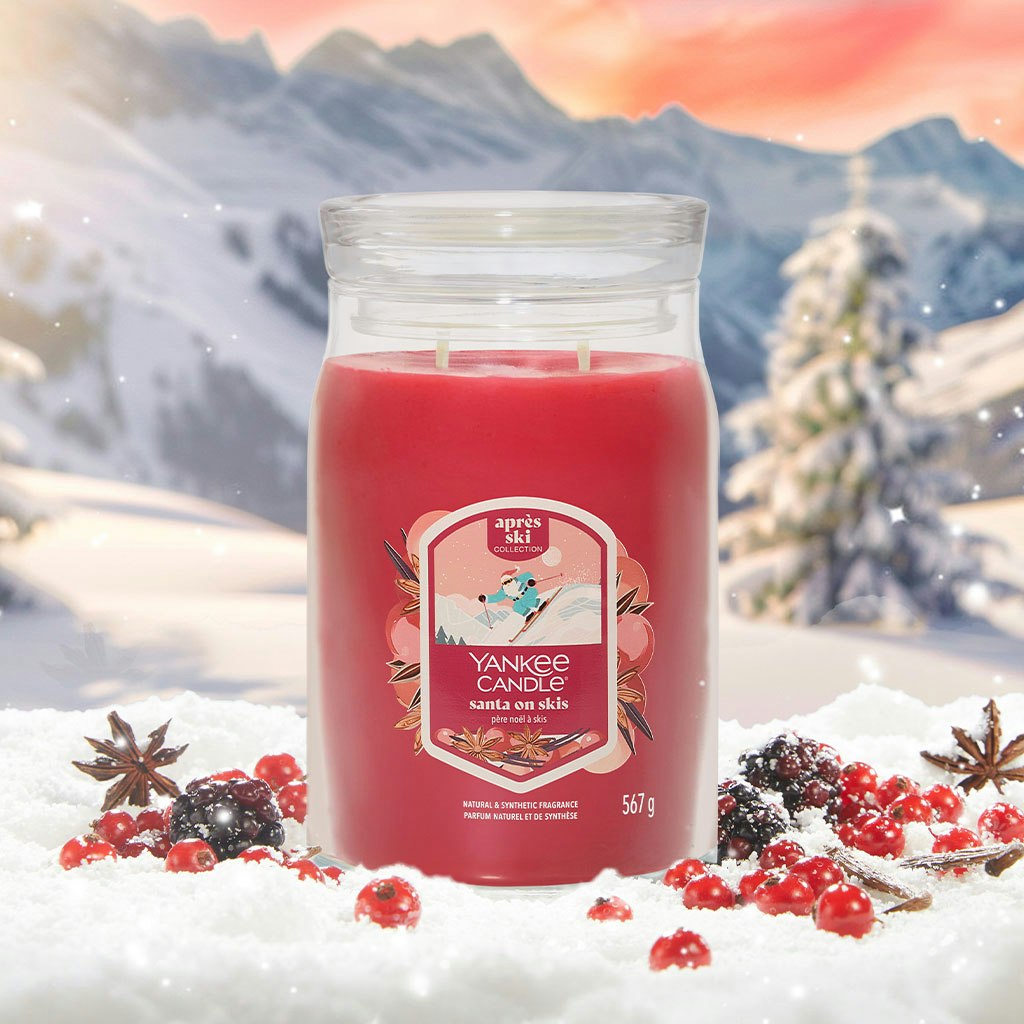 Large red holiday candle in glass jar surrounded by winter berries and snow, mountain scene in background