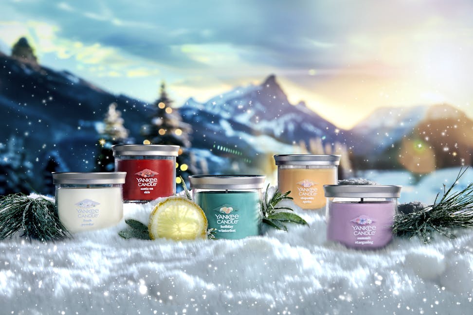 Apres Ski Collection Fresh Powder Signature Small Tumbler Candle, Santa on Skis Signature Small Tumbler Candle, Holiday Winterfest Signature Small Tumbler Candle, Slopeside Spritz Signature Small Tumbler Candle, and Summit Stargazing Signature Small Tumbler Candle in snow surrounded by branches, slices of lemon, and mountains