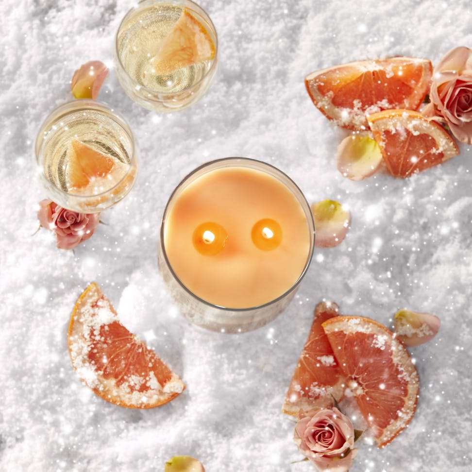 top down look at a lit two wick candle in the snow with sliced grapefruit and roses