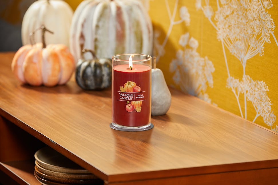 lit apple pumpkin signature medium pillar candle on wooden table next to pumpkins and yellow floral wallpaper