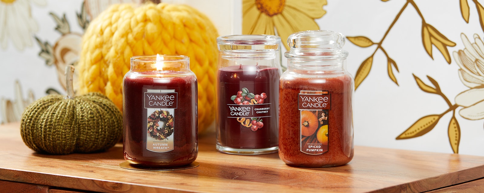 three orange and red large jar candles on table with knit pumpkins and flower artwork
