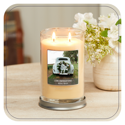 a cream-colored signature large tumbler candle with a personalized photo label of a white car and messaging reading congratulations newly weds on a wood table next to a vase filled with white flowers