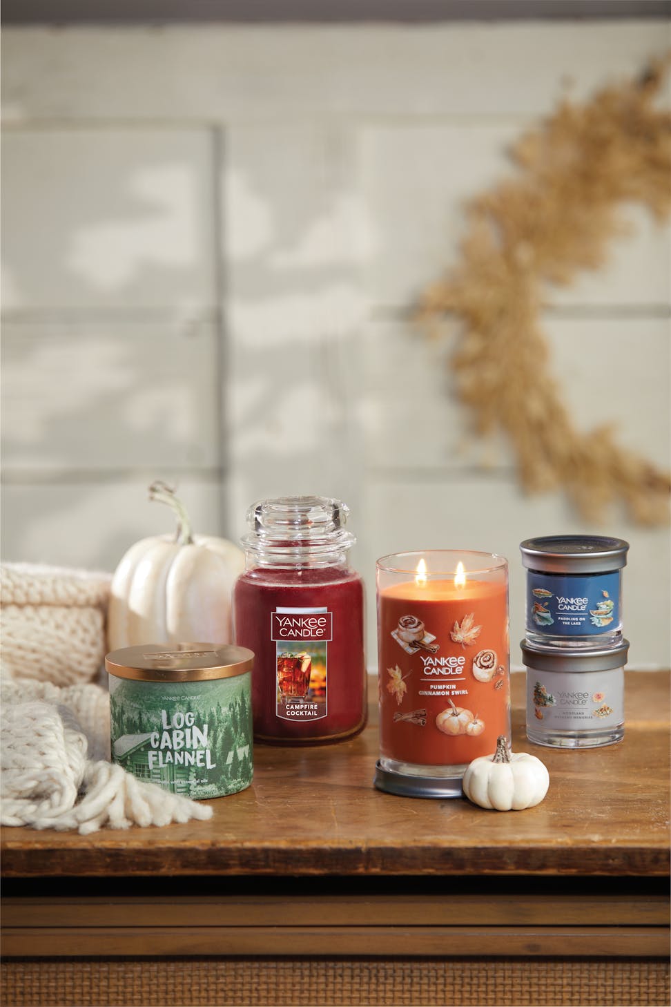 a log cabin flannel 3-Wick candle, a campfire cocktail original large jar candle, a pumpkin cinnamon swirl signature large tumbler candle, a paddling on the lake signature small tumbler candle, and a woodland weekend memories signature small tumbler candle on a table with two white pumpkins and a white blanket