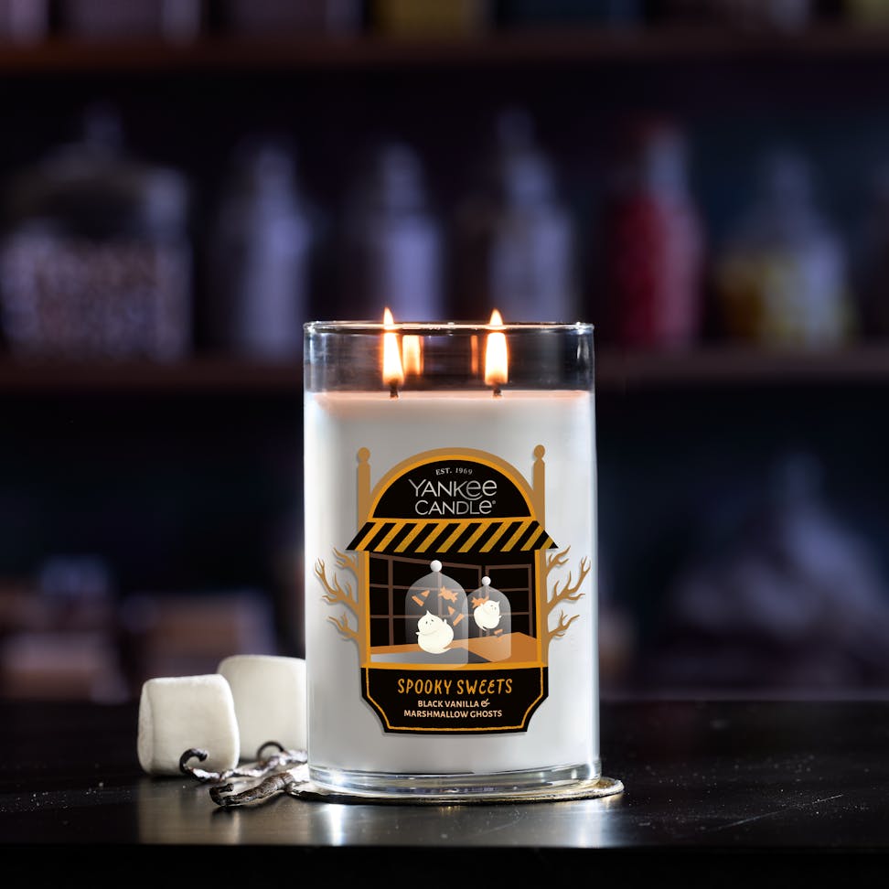 Large glass jar candle with two wicks burns beside marshmallows, bakery shop and spooky sweets label on front.