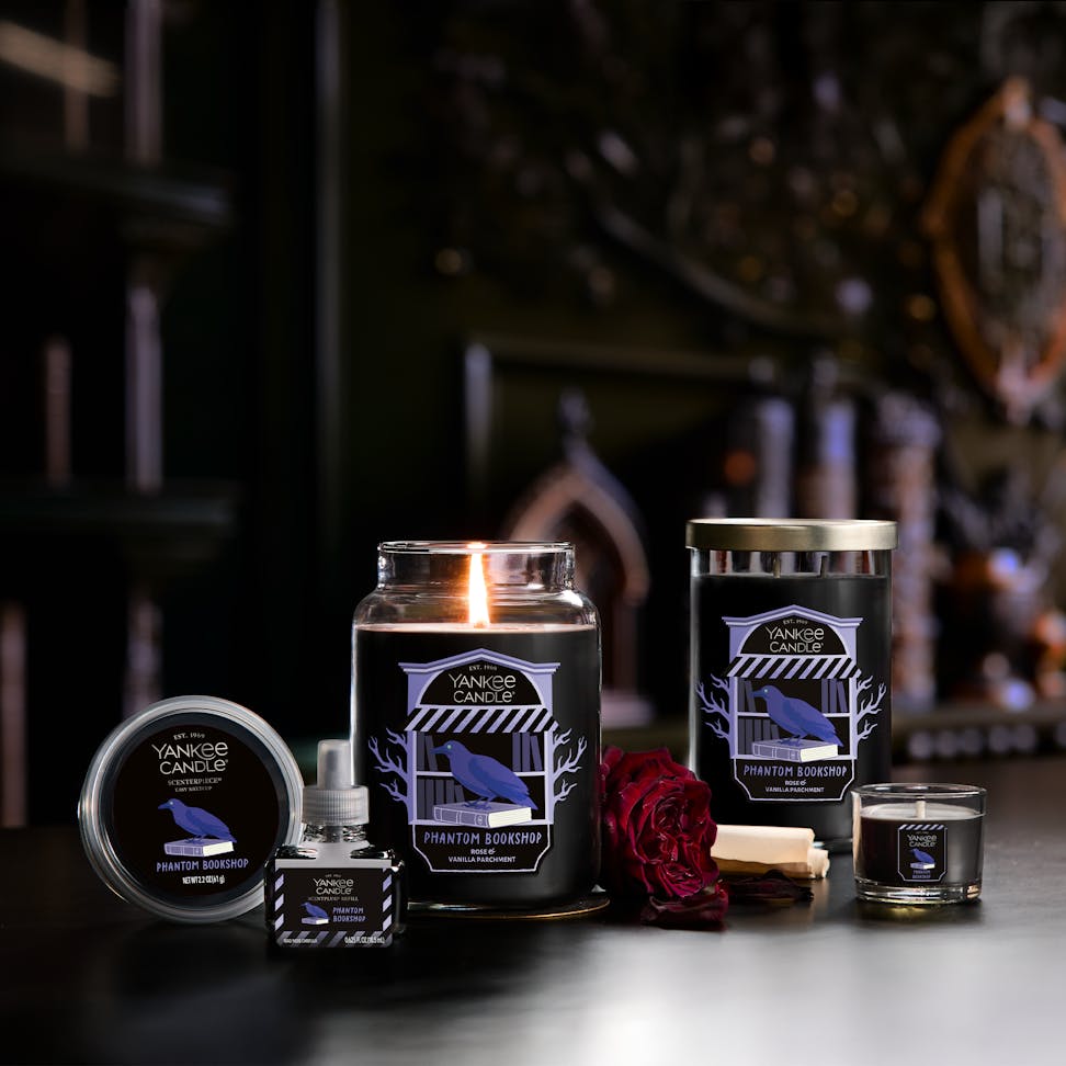 Large black jar candle with raven label, surrounded by rose, books, and other candles in a cozy home setting