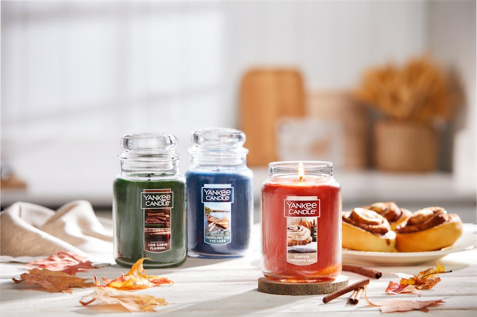 Cottage Getaway Collection Log Cabin Flannel Original Large Jar Candle, Paddling on the Lake Original Large Jar Candle, and Pumpkin Cinnamon Swirl Original Large Jar Candle on a table with cinnamon rolls, leaves, and sticks of cinnamon