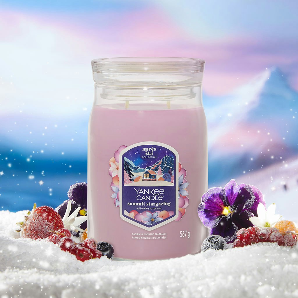 Large pink scented candle in a glass jar surrounded by snow, berries, and flowers with a snowy mountain scene