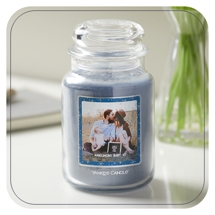 a blue-colored original large jar candle with a personalized photo label with a man, a woman, and a young child and announcing baby #2 messaging on a white table