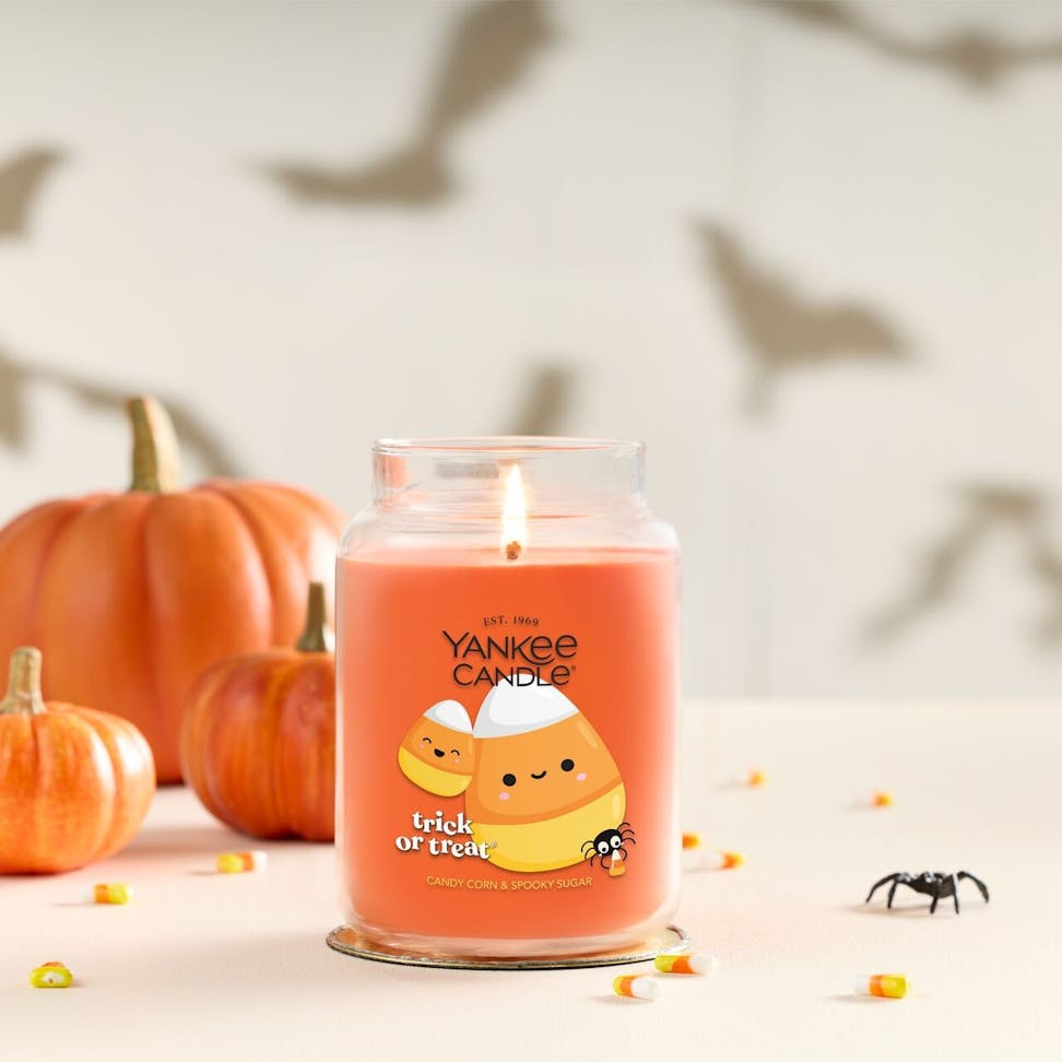 Large orange candle jar with candy corn design, surrounded by pumpkins, candy, and Halloween bat wall decor.