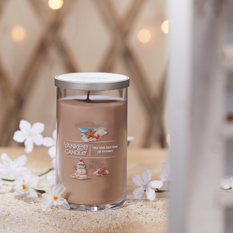 Passport to the Holidays Collection Tis the Sea-Sun in Sydney Signature Medium Pillar Candle surrounded by sand and white flowers