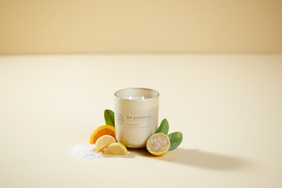 chesapeake bay candle intentions collection be positive citrus sugar medium two wick jar candle surrounded by orange slices and sugar in a pastel yellow backdrop